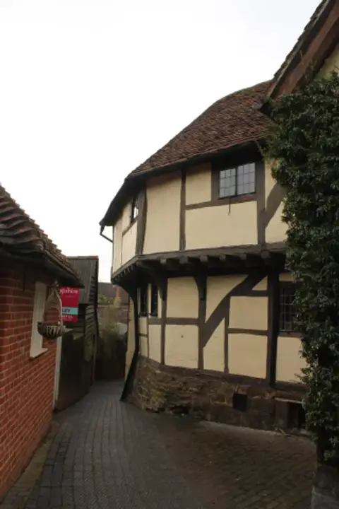 Jettied timber-framed rear elevation on a stone plinth, above a brick-paved lane