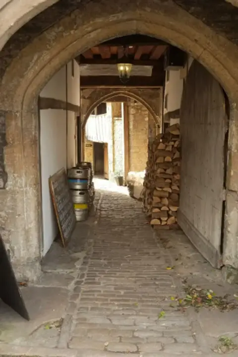 Pointed stone archway leading to a cobbled passage with beer kegs and stacked firewood