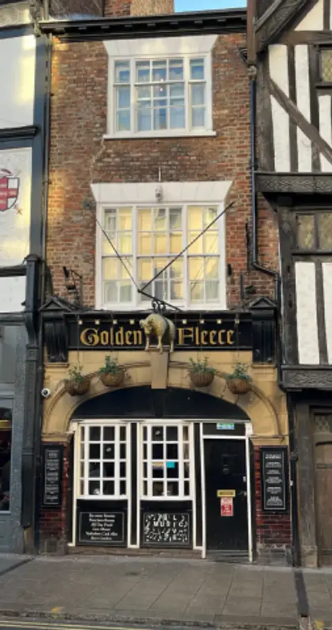 The same brick frontage in low sun, with hanging baskets and boards listing the pub's ghosts