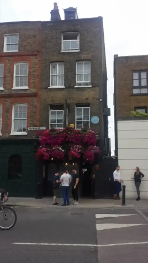 Narrow brick pub on a corner with a dark green front, a mass of pink flowers and a blue plaque