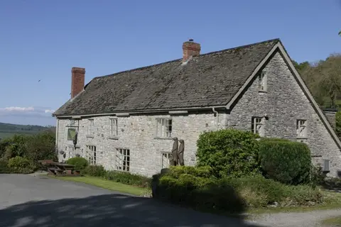 A long stone longhouse with a stone-tiled roof and two brick chimneys, standing above open hill country