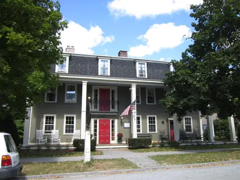 Grey clapboard inn of two storeys with a gambrel roof, dormer windows, white porch posts and red doors