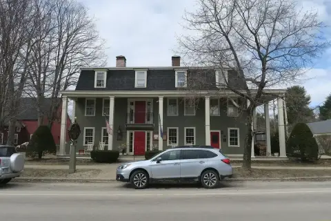 The same clapboard front in early spring, bare trees above it and a roadside sign lettered Hancock Inn
