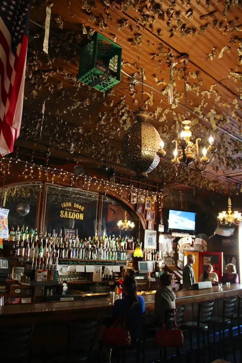 Long saloon bar under a timber ceiling covered in pinned dollar bills, with a mirrored back bar
