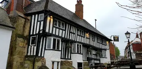 Black-and-white timber-framed inn with leaded windows and a stone plinth, beside a flint wall
