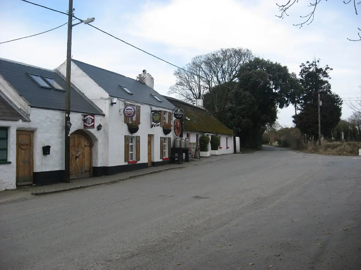 White roadside inn with a thatched range beyond, Guinness signs and a cartwheel on the wall