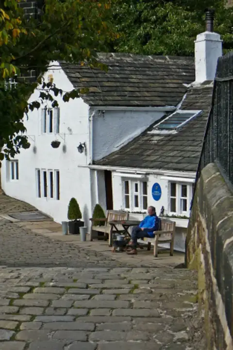 Whitewashed inn with stone-slate roof at the foot of a cobbled lane, benches by the door