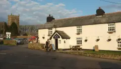 Long white rendered inn with a slate roof and hanging sign, the granite church tower behind