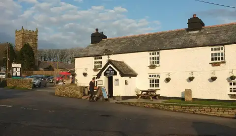 Long white rendered inn with a slate roof and hanging sign, the granite church tower behind