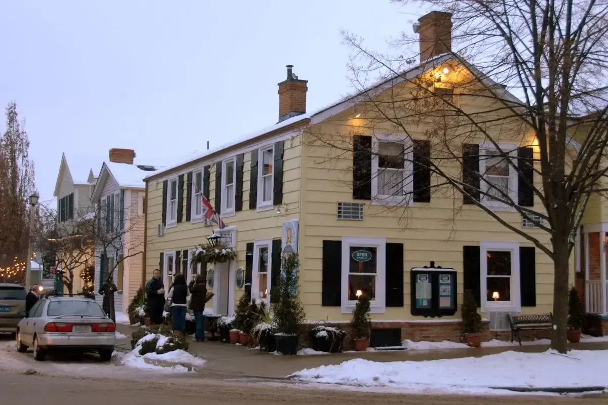 Pale yellow clapboard inn with black shutters and brick chimneys, snow along the pavement