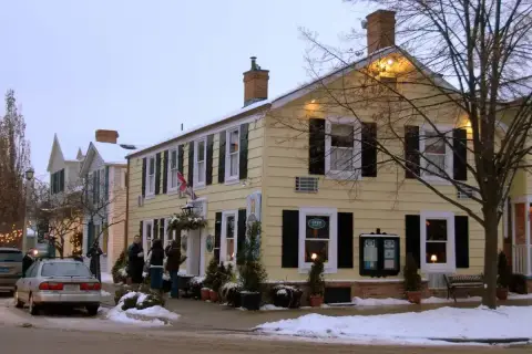 Pale yellow clapboard inn with black shutters and brick chimneys, snow along the pavement
