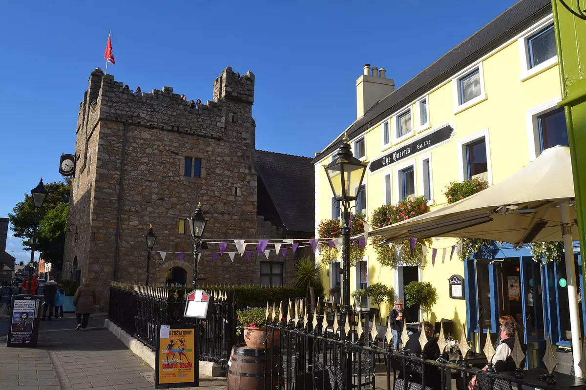 Yellow three-storey pub with a black The Queen’s Est. 1745 fascia and flower boxes, beside a stone castle