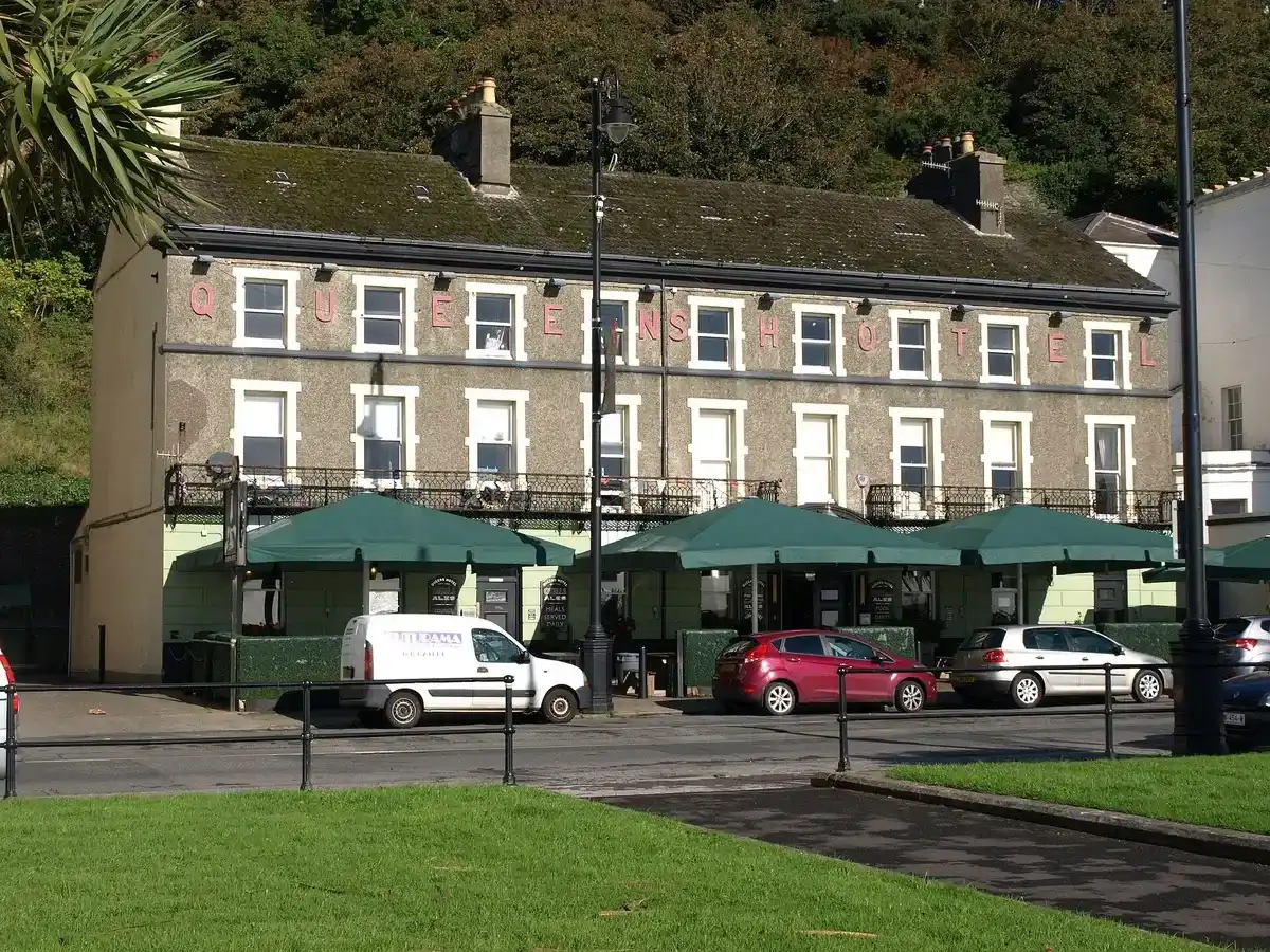 Three-storey stone pub with QUEENS HOTEL in raised pink letters and green parasols along its front