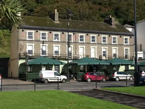 Three-storey stone pub with QUEENS HOTEL in raised pink letters and green parasols along its front