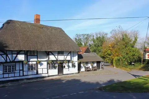 Deep thatched roof and white panels between black timbers, with a brick chimney and hanging baskets