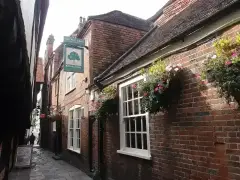 Brick pub wall with sash windows and flower baskets along a narrow passage, hanging oak-tree sign