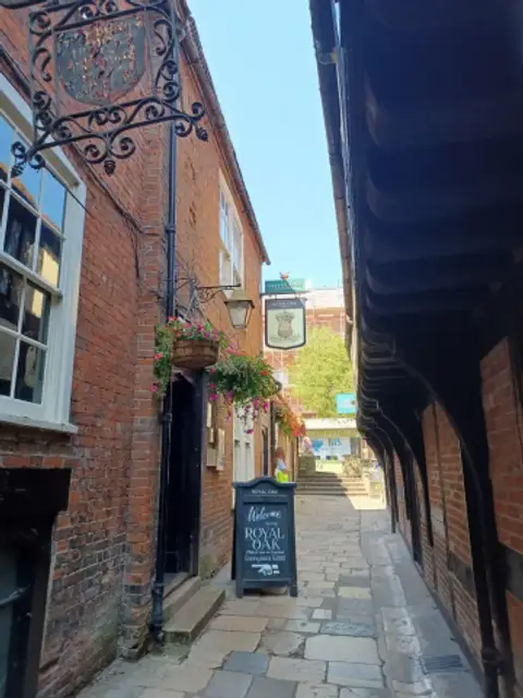 Narrow flagstone passage between brick and timber buildings, with a pub doorway and A-board