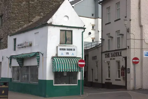 Pub's cream corner elevation lettered The Saddle, est. 1843, beside a closed green and white shop