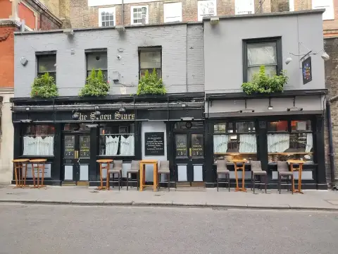 Black-painted pub front with gold lettering, window boxes above and high stools on the pavement
