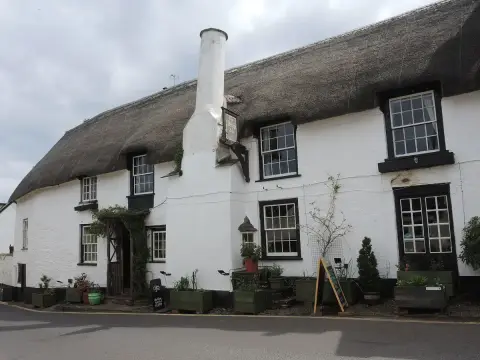 Long white thatched inn with black window frames and a hanging sign, seen from the lane