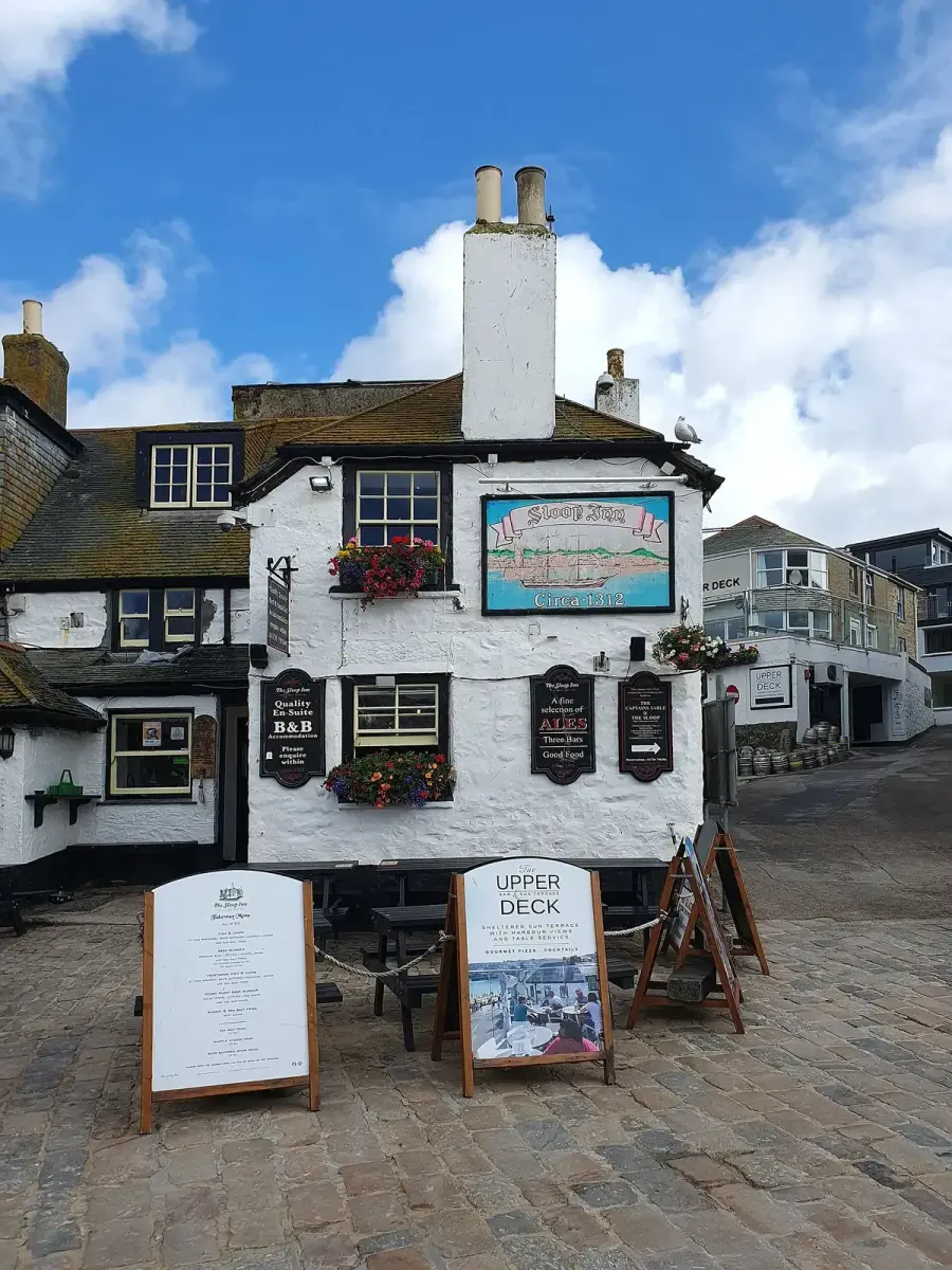 Whitewashed harbour inn with a tall chimney and painted sloop sign, on the cobbled wharf