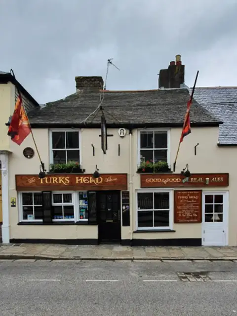Cream-painted pub front with a carved timber fascia, slate roof and two flags on poles