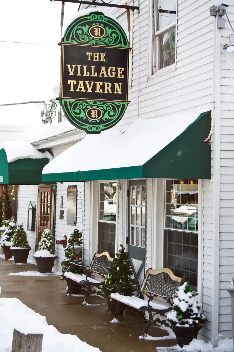 White clapboard front of the Village Tavern in snow, with its oval hanging sign, green awnings and benches along the pavement