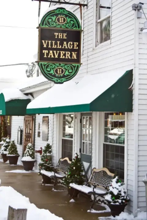 White clapboard front of the Village Tavern in snow, with its oval hanging sign, green awnings and benches along the pavement