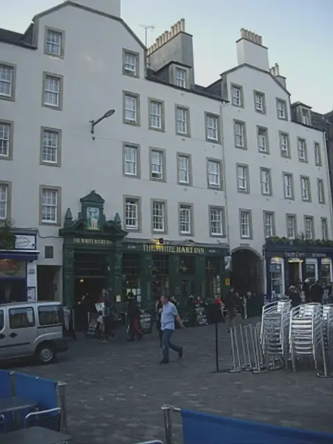 The green pub front seen across the Grassmarket setts, in a row of tall harled tenements
