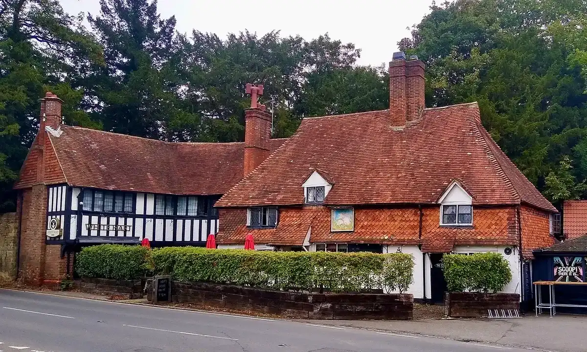 Long tile-hung and timber-framed pub behind a clipped hedge, brick stacks and red parasols