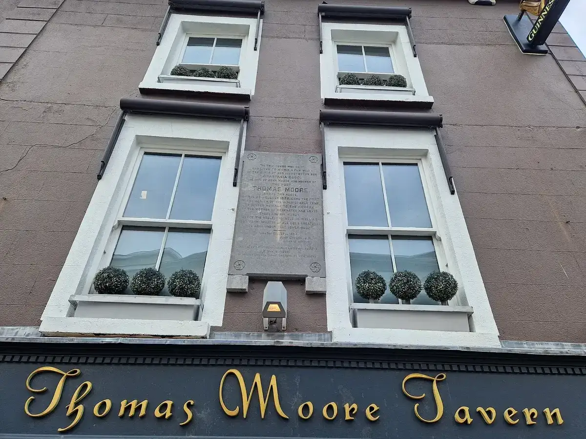 A tavern front in gold script lettering reading Thomas Moore Tavern, with a stone plaque set in the wall above it between sash windows
