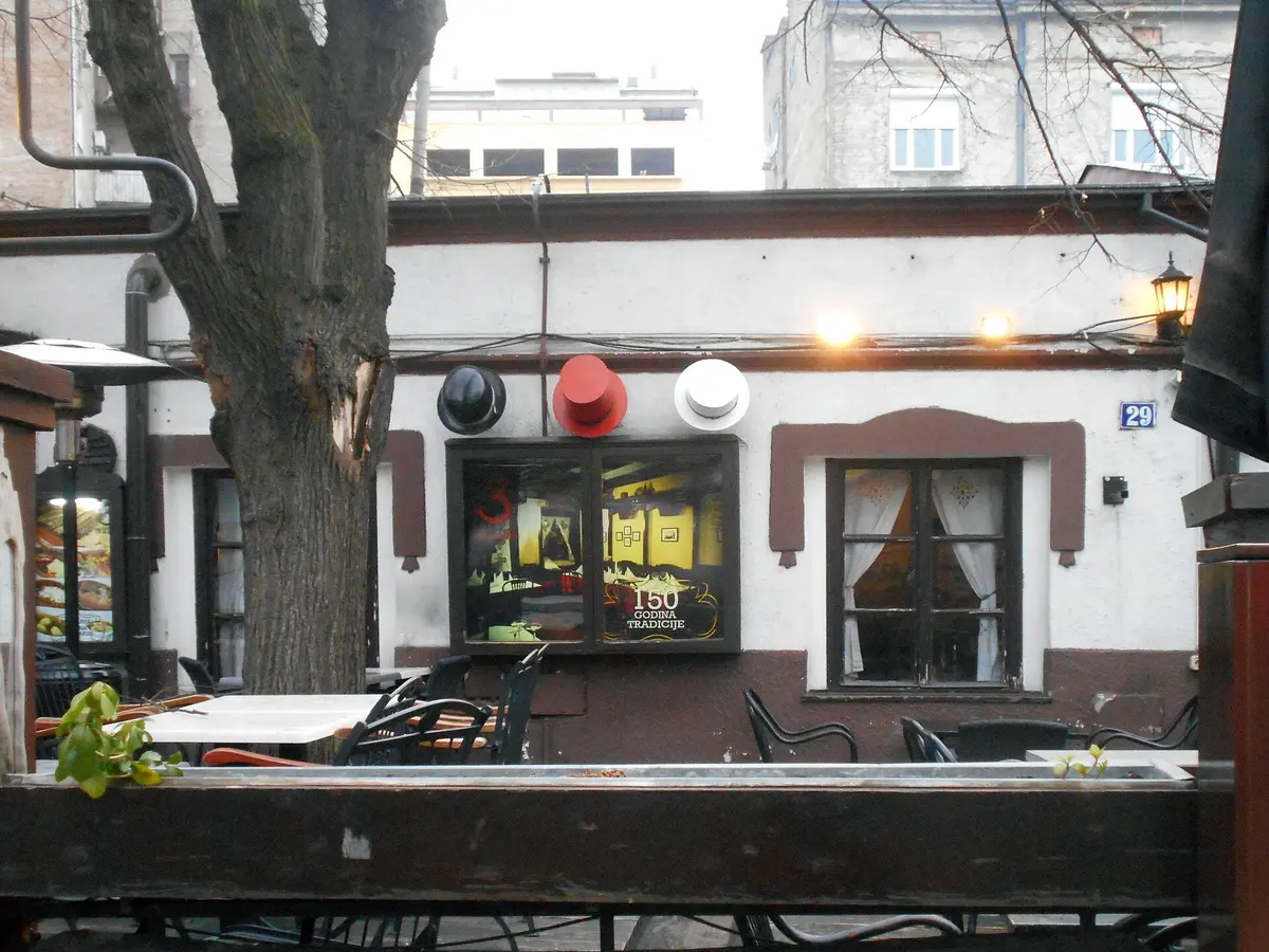 White single-storey kafana at number 29 with black, red and white hats mounted above a window
