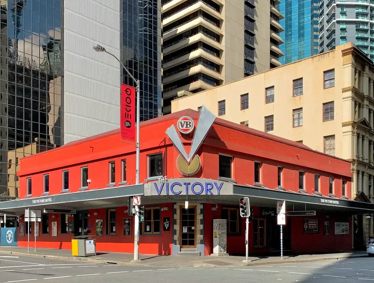 Red-painted corner hotel with a wide awning lettered The Victory, office towers rising behind it