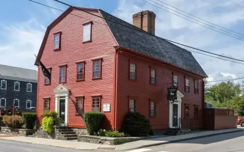 Dark red clapboard tavern with a gambrel roof, a huge brick chimney and white pedimented doors