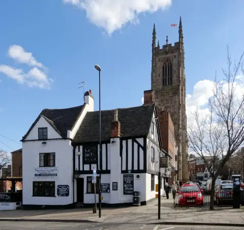 Low white and black timber-framed pub below a tall church tower, board reading Derbys Oldest Pub