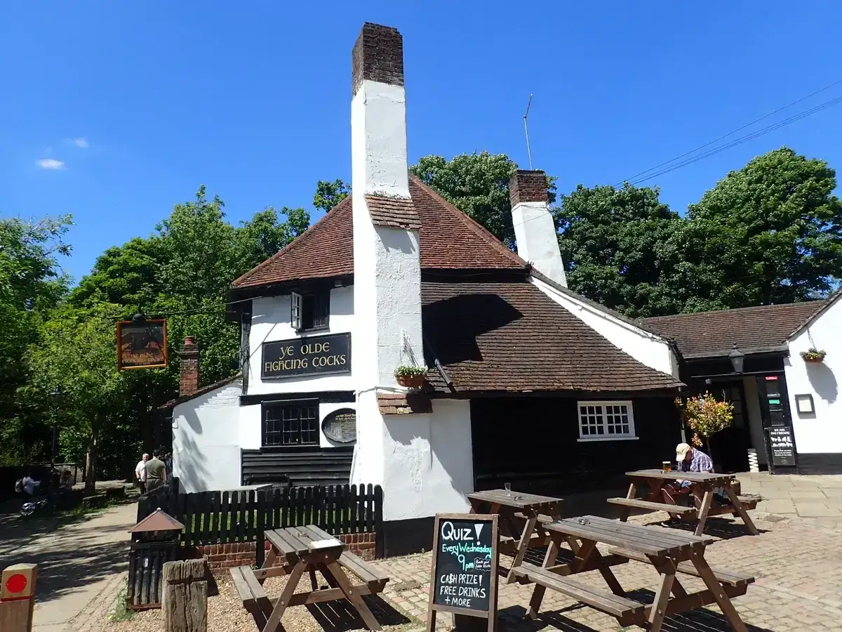 Low white-walled pub with a steep tiled roof, tall chimneys and picnic benches on the gravel outside