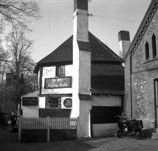 The Fighting Cocks in 1956: octagonal tiled roof, tall white chimney stack and a motorcycle parked by the fence