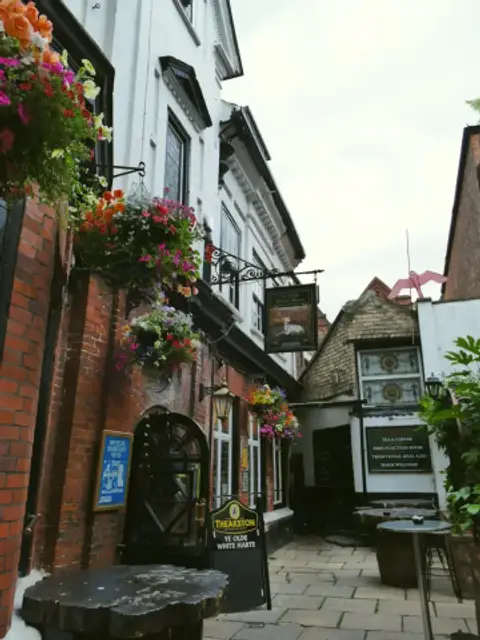 Brick and white-painted walls down a narrow flagged courtyard, with hanging baskets and a pub sign