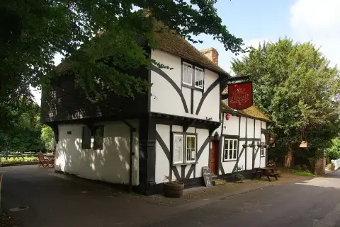 Small jettied timber-framed inn under a tiled roof with a red hanging sign, dark yew behind