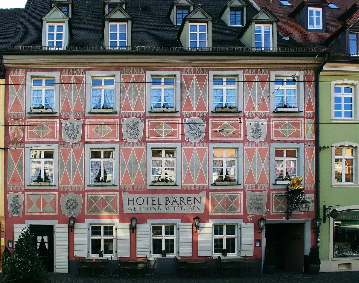 Pink and white painted front with coats of arms and dates, lettered Hotel Bären, tables set out below