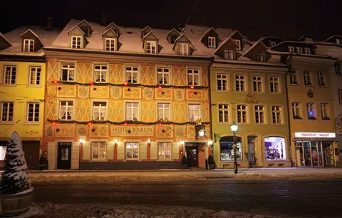 The same painted front at night in snow, lamps lit below the windows and dormers along the roof