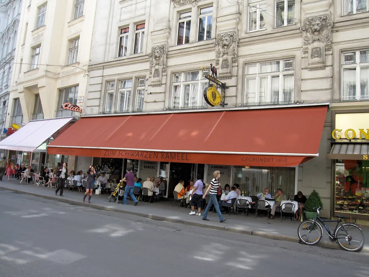 Red awning lettered Zum Schwarzen Kameel gegründet 1618 over pavement tables, camel sign above